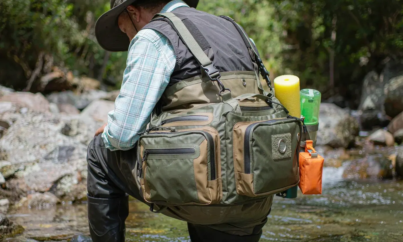 Angler in waders with a fishing waist pack and a sling pack rotated forward for tray access in shallow water