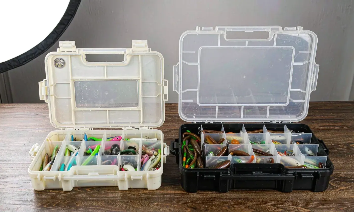 Two fishing tackle boxes side by side showing the size difference between a 3600 and a larger 3700 tray resting on a sloping riverbank